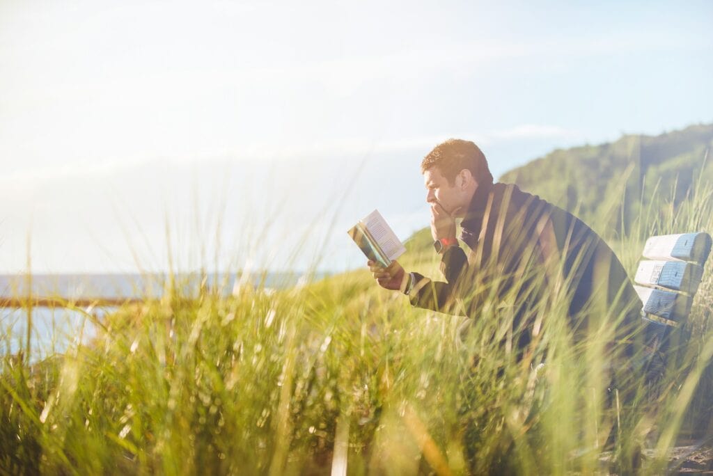 An adult man reads a book in a meadow.